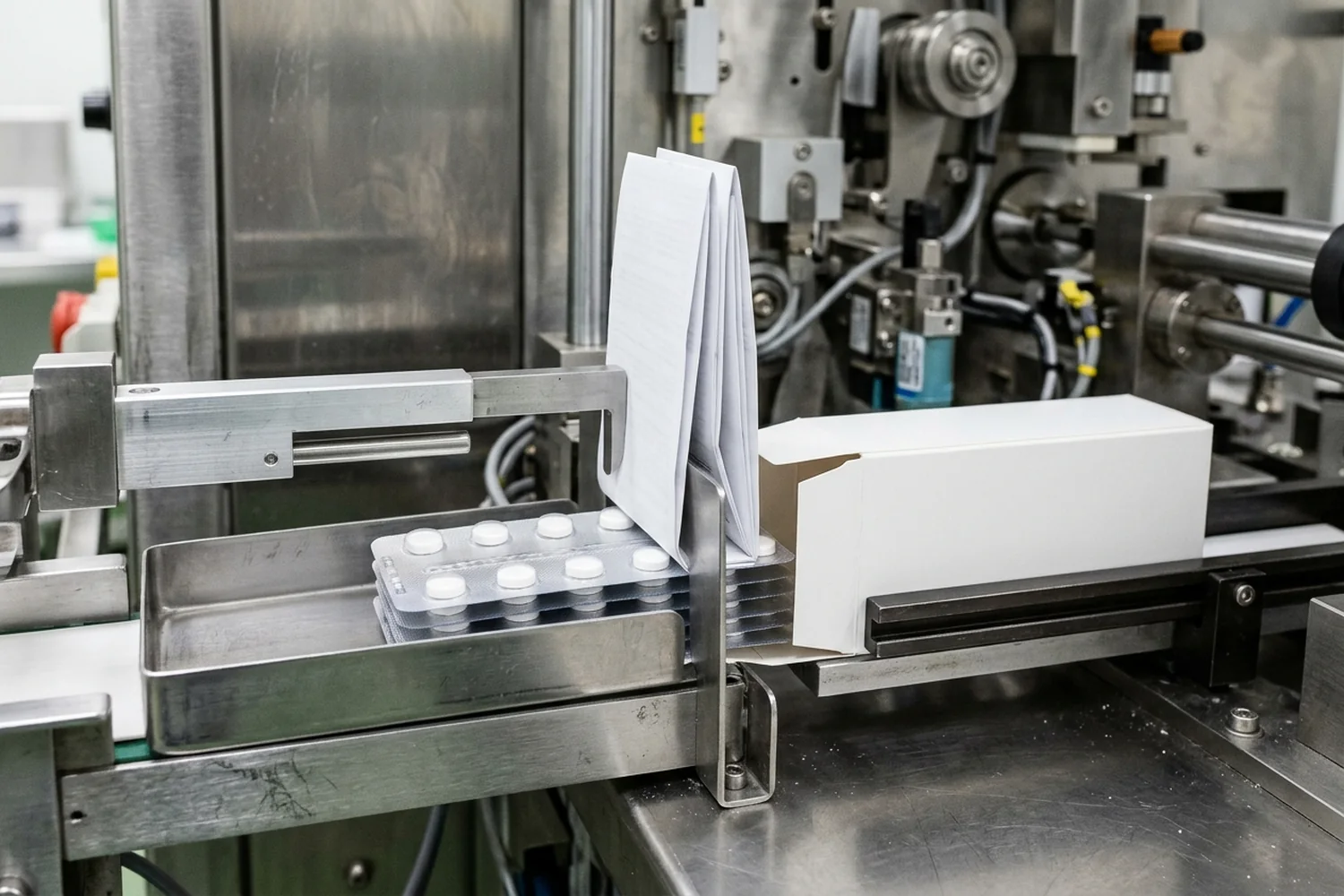 Stacked tablet blister cards inside a stainless-steel bucket guide advancing toward a folded white leaflet held vertically across an open horizontal carton sleeve